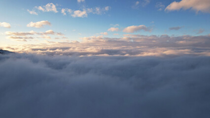 Flying among the ocean of clouds in the mountains. The fog is like big ocean waves crashing against high mountains covered with snow. Spruce trees grow in places. Sunset. Orange rays of the sun