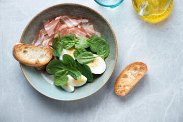 Plate with fresh spinach leaves, bacon, hard-boiled eggs and ciabatta, above view on a light-grey stone background, horizontal shot