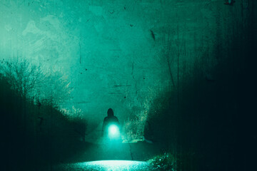 A scary hooded figure carrying a torch, silhouetted along a mysterious country road. On a spooky foggy winters night. With a grunge edit.