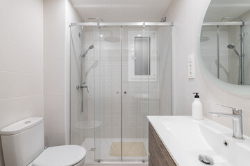 Spacious white ceramic tile bathroom is illuminated by bright white natural light. Large round mirror on the wall reflects the shower area with glass railing and metal fittings.