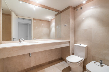 Fototapeta na wymiar Modern spacious bathroom with bright beige tiles, toilet, bidet and sink on long marble countertop. Full-length mirror reflects open door to hallway. Bright light from bulbs on ceiling.