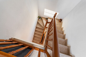 View from above of spacious, daylight-lit spiral staircase with beige marble steps and wooden railings leading down from top floor. On steps there are pots with indoor plants for home comfort.