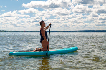 A woman in a sports swimsuit with a paddle on her knees on a SUP board in a lake against the background of white clouds on a clear blue sky.