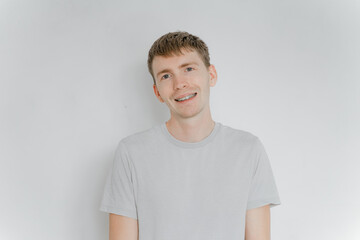 a man in a gray t-shirt on a white wall stands smiling with braces