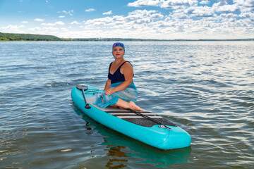 A woman with a Mohawk in a pareo on a SUP board in a lake against the background of white clouds on a clear blue sky.