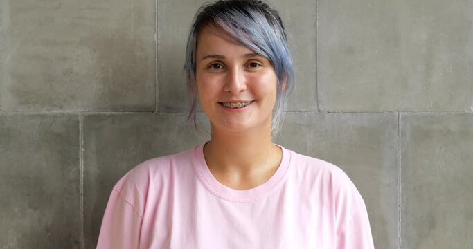 Portrait shot of young woman with casual clothes and blue hair. She look straight then smile, we see dental braces on her teeth. Then she burst out laughing and shy little, tilt down head and body