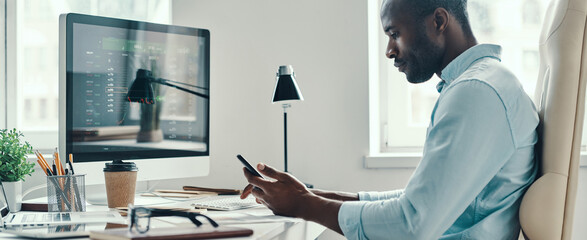 Thoughtful young African man in shirt using smart phone while working in the office
