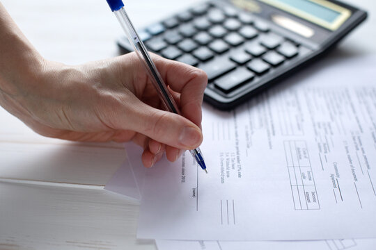 A photo of a person's hand holding a pen and filling out papers. Calculator in the background