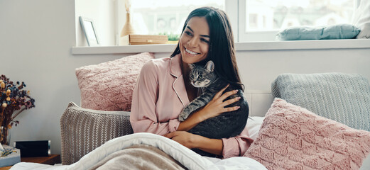 Attractive young woman in pajamas carrying a cat and smiling while resting in bed at home