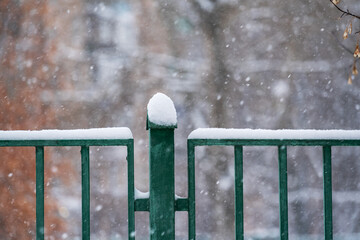 A snow-covered green fence in close-up against the background of multi-storey buildings. Snow sweeps all surfaces on the street