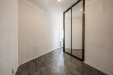 Obraz na płótnie Canvas An empty room with a built-in wardrobe with sliding mirror doors with ocher anodized aluminum edges, white painted walls with rough gotelet and gray wooden floors