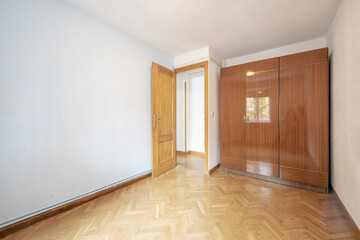 An empty room with a wardrobe with three-section glossy veneered doors with oak parquet floors placed in a herringbone pattern and hot water pipes on the skirting board