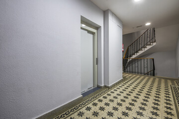 Landing of an urban residential housing building with stairs with wrought iron railings, hydraulic tiled floors and an elevator with light metal doors
