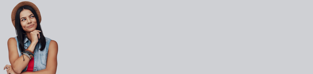 Thoughtful young woman in hat holding hand on chin while standing against grey background