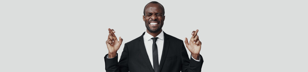 Handsome young African man in formalwear making a face and keeping fingers crossed while standing against grey background
