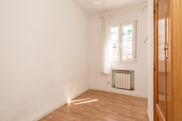 An empty room with an aluminum radiator within a niche below a double-hung window and an oak built-in cupboard