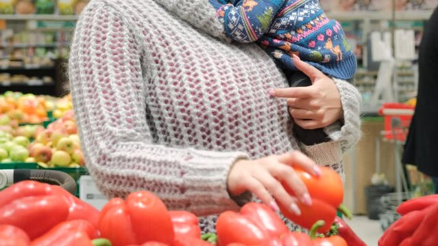 Close-up of young woman holding baby in hands and buying bell peppers at grocery store