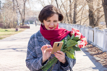 a woman with a bouquet of beautiful roses is texting on the phone in the autumn city park. clear sunny day