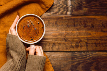 Woman hands in sweater with cup of coffee on linen towel on dark wooden background, copy space for product presentation