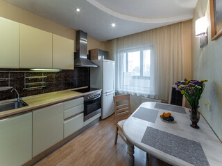 Modern interior of kitchen in apartment. Table and chairs. Fridge, oven and fan. Window with tulle.