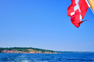 Flying Danish flag at the stern of an excursion boat and picturesque view of the coastline of Gudhjem, Bornholm island, Denmark.