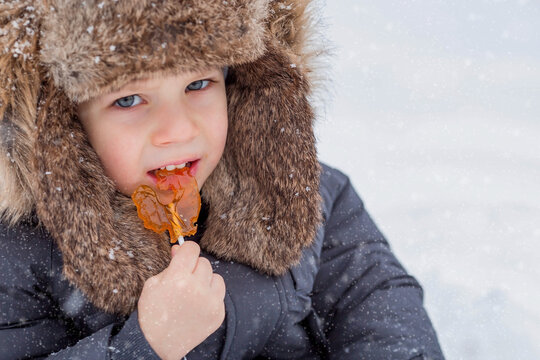 Portrait of a beautiful little Russian boy in a hat with earflaps with a lollipop cockerel in winter in the park. The concept of a happy childhood, Russian traditional sweets and lifestyle.