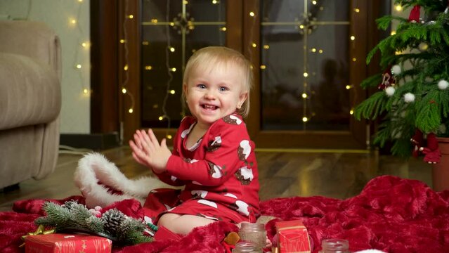 A sweet girl of one and a half years claps her hands. Garlands and a Christmas tree. Christmas and New Year and children