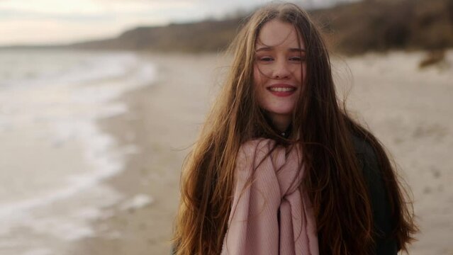 Happy mixed ethnic girl smiling and looking at camera on sea shore. Portrait of loving joyful asian woman laughing, standing on ocean coast. Wind blows her airy hair. Dreamy cinematic slow motion.