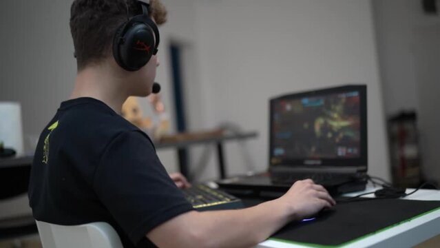 A teenage guy playing a computer game on a laptop. The gamer sits on a chair wearing headphones with a microphone and quickly presses the buttons of the keyboard and moves the mouse. The boy