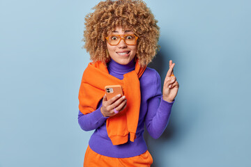 Hopeful curly haired woman bites lips uses mobile phone makes wish keeps fingers crossed bites lips wears spectacles purple turtleneck orange jumper tied over shoulders isolated over blue background