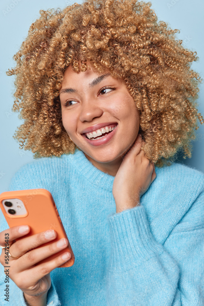 Canvas Prints Portrait of cheerful curly haired woman smiles broadly looks pensively away uses mobile phone for online communication dressed in casual soft jumper isolated over blue background. Modern technologies