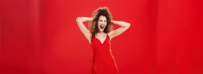 Girl just wanna have fun. Crazy adult caucasian woman with curly hairstyle in elegant dress holding hands on head yelling out loud with closed eyes and intense expression over red background - obrazy, fototapety, plakaty