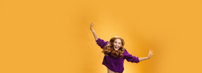 Fototapeta na wymiar Expressive and enthusiastic carefree friendly joyful redhead woman in purple sweater and pants jumping joyfully with raised hands smiling yelling from happiness and joy against orange background
