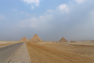 Fototapeta na wymiar The three great pyramids of the Giza complex, seen from the back, with the yellow sand, a road next to the pyramids and the sky with clouds.