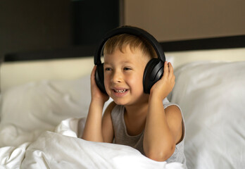 Little sleepy boy sitting in bed wearing wireless headphones and listening to music