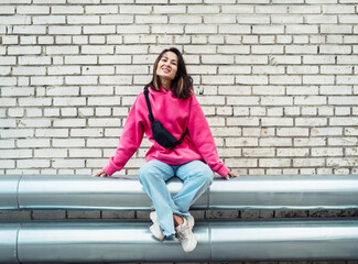 Fashion Asian young woman in a pink sweatshirt and jeans sits against a brick wall background, has...