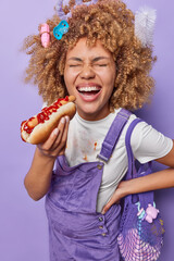 Nutrition during pregnancy. Joyful pregnant woman wears white dirty t shirt and overalls eats delicious hot dog with ketchup carries net bag on shoulder laughs happily isolated over purple background