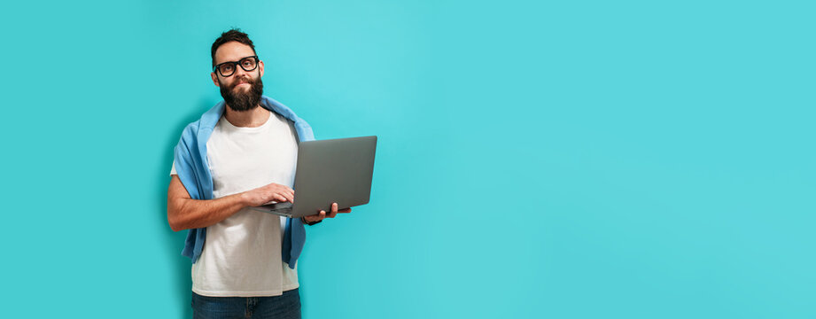 Young bearded charismatic man hipster. Shocked or surprised expression. Laptop concept. Promotion poster. Programmer, web developer holding a laptop in his hands and looking at the camera