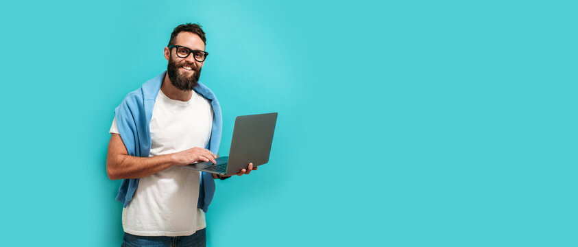 Young bearded charismatic man hipster. Shocked or surprised expression. Laptop concept. Promotion poster. Programmer, web developer holding a laptop in his hands and looking at the camera