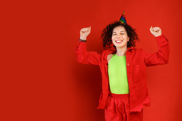 Curly cute girl laughs and jumps in a colorful birthday hat. Studio portrait of a woman on a bright red background. Positive smiling young female.