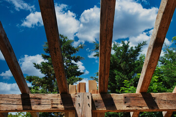 An incomplete wooden roof skeleton in nature with sky and clouds frame, tree branches in the sky. Roof repairing.