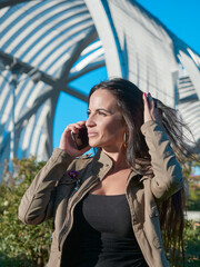 Woman long brown, brown hair. Woman next to a bridge talking on mobile phone. Blue sky and green...