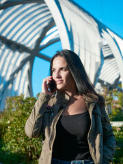 Woman long brown, brown hair. Woman next to a bridge talking on mobile phone. Blue sky and green park. Focus selection. Metal arches