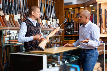 Confident experienced salesperson helping Hispanic customer choose right hunting rifle at gun store - obrazy, fototapety, plakaty
