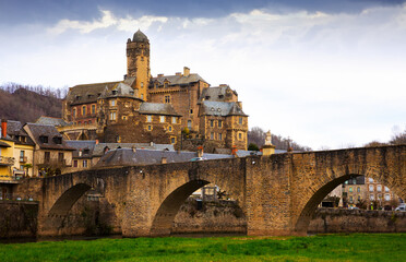 View of medieval castle of Chateau Estaing and old arched bridge across river Lot in cloudy winter...