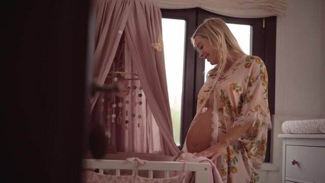 Exited young pregnant woman contemplating her baby crib and daydreaming smiling. Maternity-wear showing bare belly.