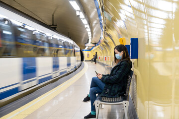 Young latin woman seated and wearing surgical mask with smartphone while waiting in the metro or...