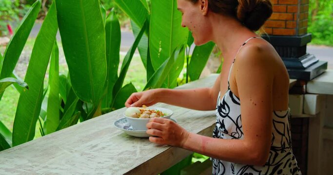 Girl eating delicious traditional balinese bakso ayam soup outdoor with lush green nature background. Woman enjoying lunch Meatball Chicken Noodles served in bowl. Nusa Penida Bali Island Indonesia.