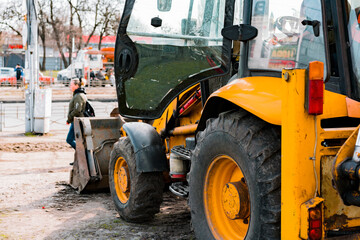 Yellow excavator stands on the ground near a sidewalk. Repair in the city. Preparation. Machine. Municipal work. Backhoe parked and ready for use for road construction on suburban street