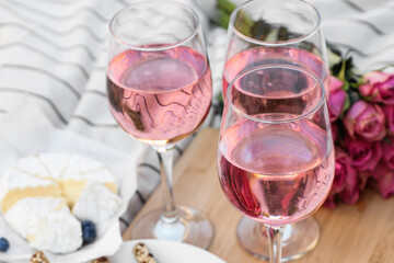 Glasses of delicious rose wine, flowers and food on white picnic blanket, closeup